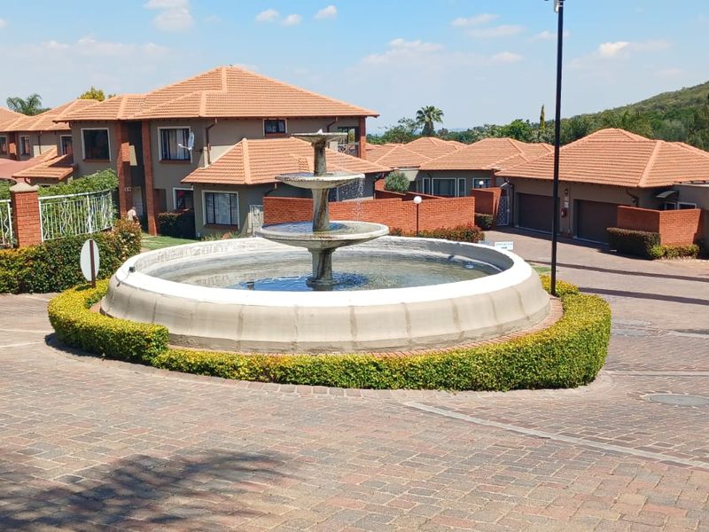A view of the townhouse complex entrance featuring a central water fountain, paved driveway, and well-maintained brick residential units with tiled roofs.