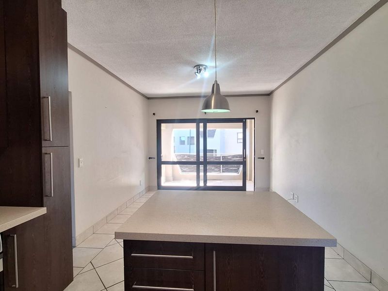 An open-plan kitchen area featuring a central island, dark wood cabinetry, built-in appliances, and tiled flooring, leading out to a balcony through sliding doors.