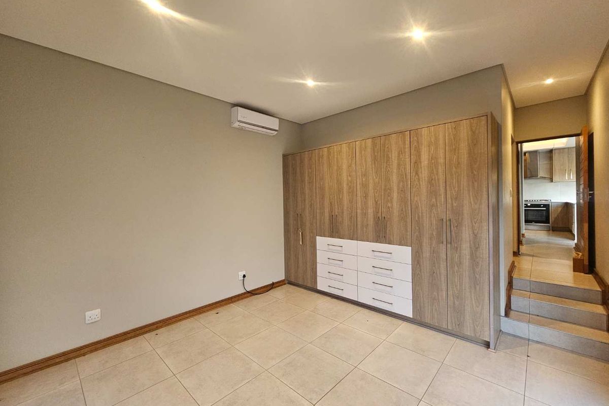 A spacious bedroom featuring large built-in wooden wardrobes with integrated drawers, tiled flooring, recessed downlighting, and a wall-mounted air conditioning unit.