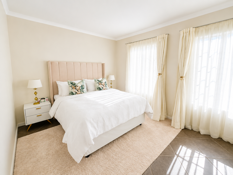 A bright and well-maintained bedroom featuring neutral wall tones, tiled flooring, and large windows dressed with elegant curtains.