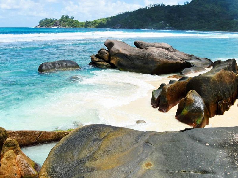 A scenic view of a pristine tropical beach with turquoise waters, white sand, and large granite rock formations.