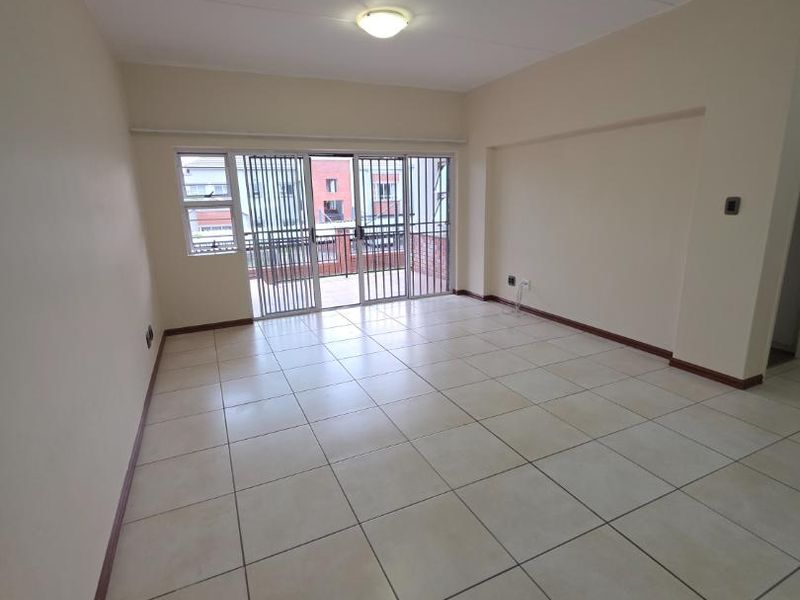 An empty, open-plan living area featuring neutral ceramic floor tiles, a sliding glass door with a security gate, and access to a balcony.