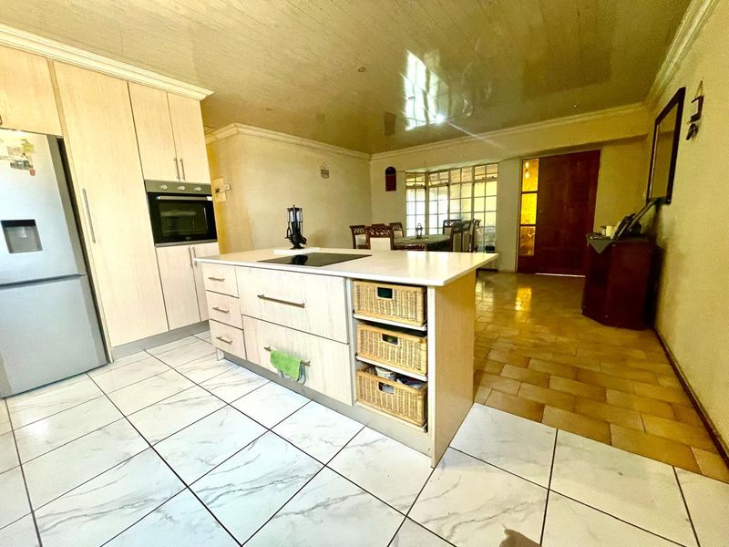 Open-plan kitchen and living area featuring a central island with integrated hob, light wood cabinetry, and contrasting tiled flooring throughout the space.