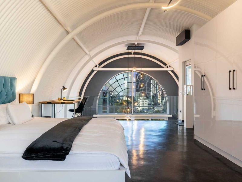 Modern bedroom featuring a unique arched corrugated ceiling, polished concrete floors, extensive built-in cabinetry, and a large arched window with city views.