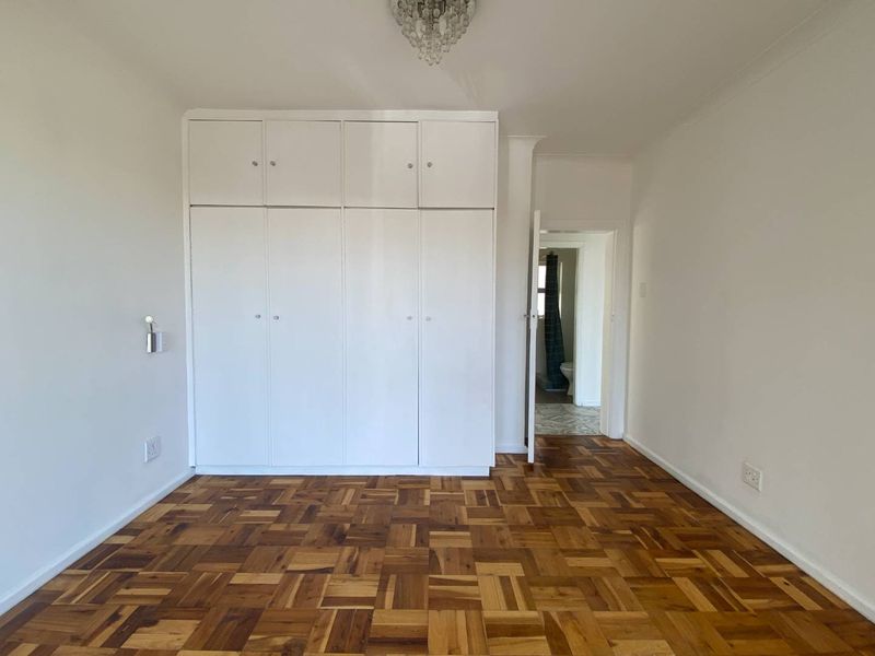 A bright, empty bedroom featuring classic parquet flooring, ample built-in cupboard storage, and a decorative crystal chandelier, with an adjacent view into the bathroom.
