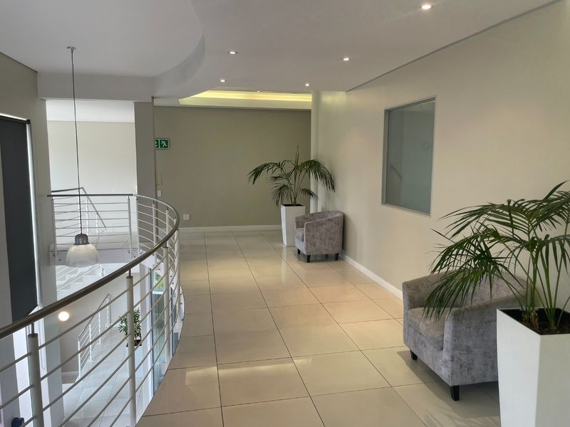 A bright and modern office hallway featuring tiled flooring, recessed downlighting, and a sleek stainless steel balustrade overlooking the lower level.