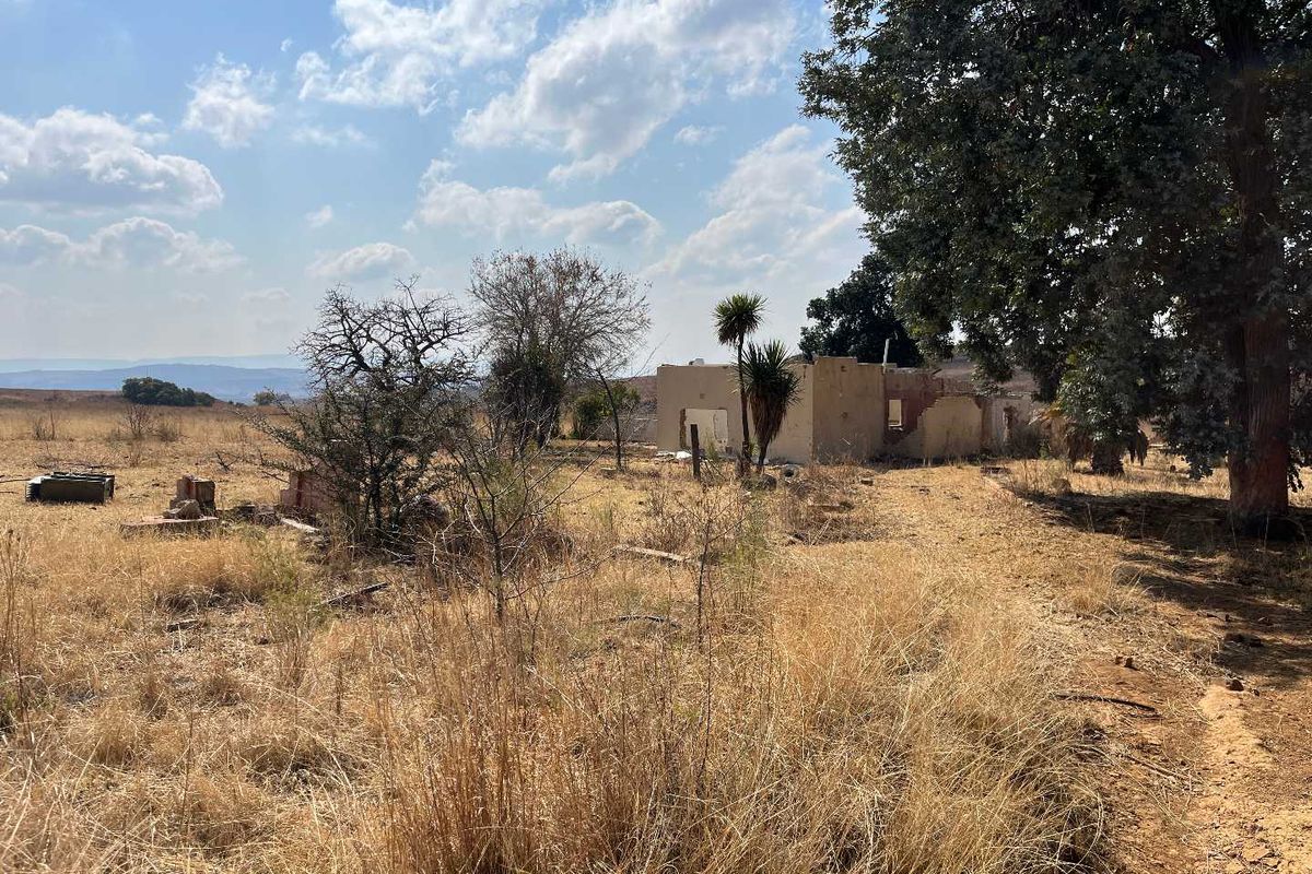 A view of a vacant agricultural plot featuring a dilapidated, partially ruined structure amidst dry, overgrown grass and mature trees.
