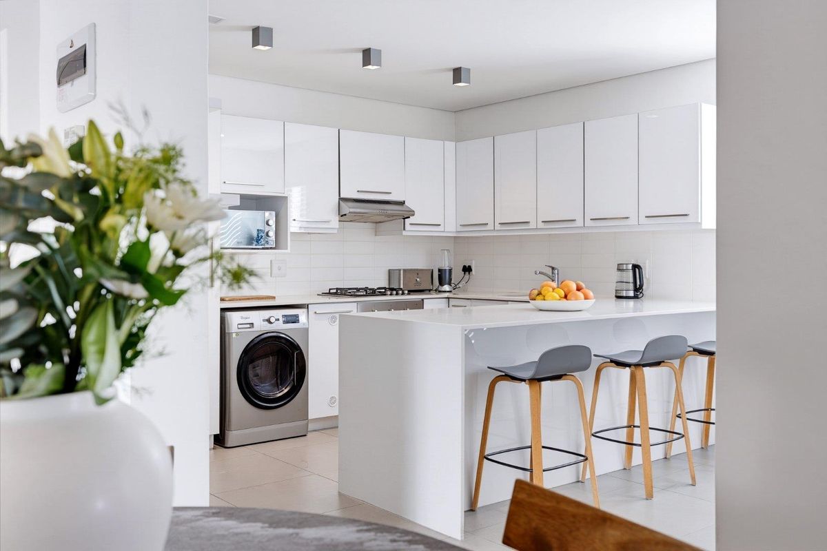 Modern kitchen featuring sleek white cabinetry, stone countertops, integrated appliances, and a breakfast bar, finished with clean tiled surfaces and contemporary lighting.