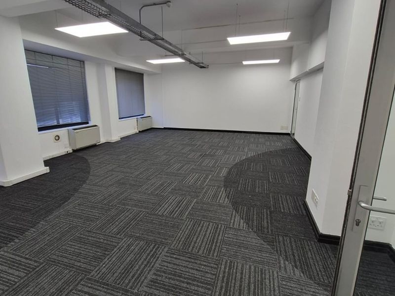 A spacious, empty office area featuring modern carpet tiles, suspended ceiling with recessed lighting, and large windows with fitted blinds.