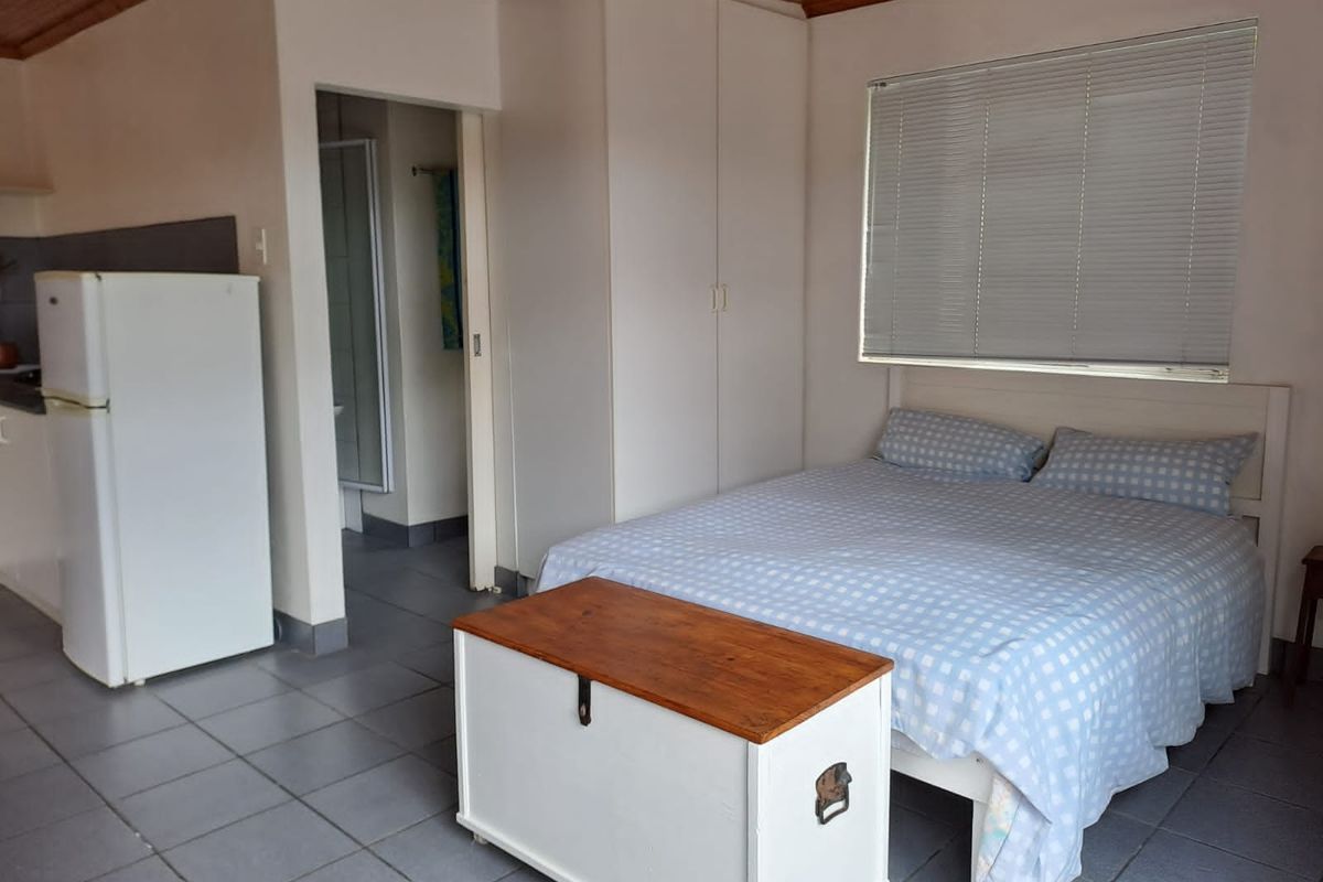 A functional bedroom featuring built-in cupboards, tiled flooring, and a timber ceiling, with an adjacent bathroom and kitchen area visible.