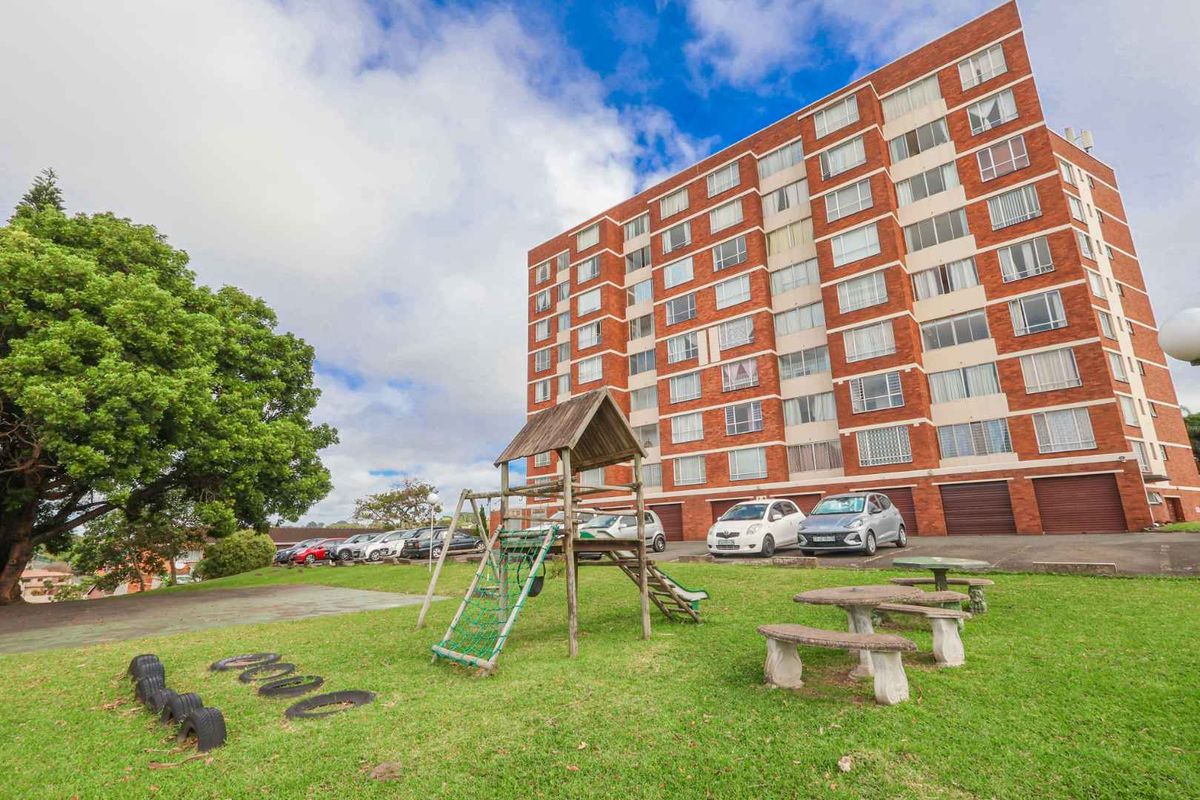 Exterior view of a multi-story brick apartment building featuring a communal garden area with a playground and parking facilities.