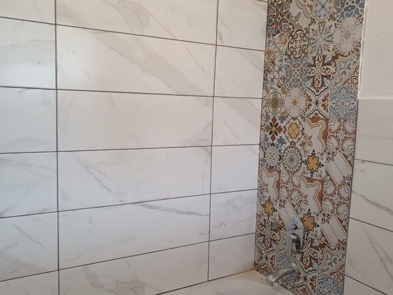 A bathroom featuring a bathtub with a shower head, finished with white marble-look wall tiles and a decorative patterned accent tile strip.