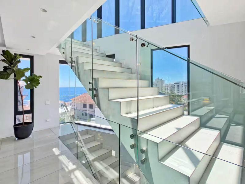 A bright, modern hallway featuring a sleek staircase with a glass balustrade, large windows, and polished tiled flooring with expansive coastal views.