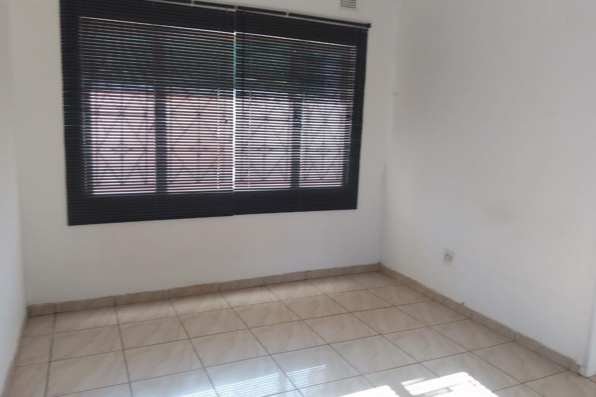 A bright, empty bedroom featuring neutral floor tiles, white walls, and dark horizontal window blinds.