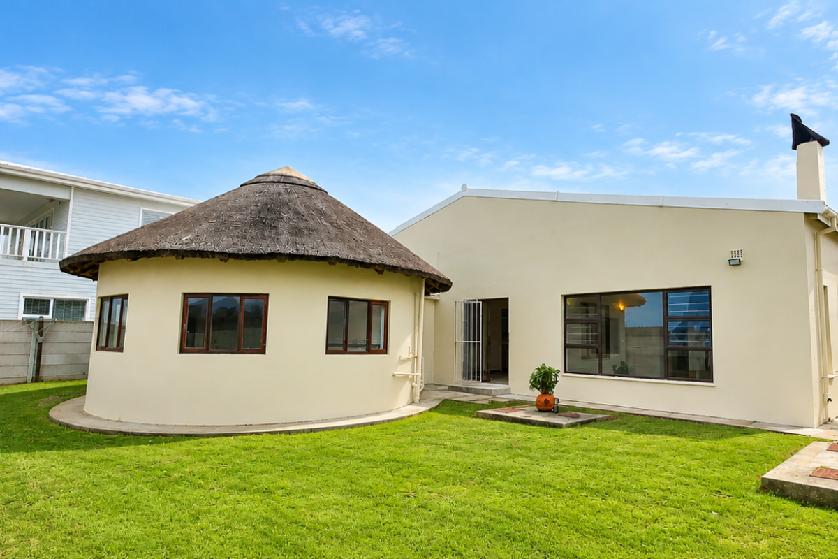 Rear view of the property featuring a unique circular thatched roof section, a spacious lawn, and perimeter boundary walls.