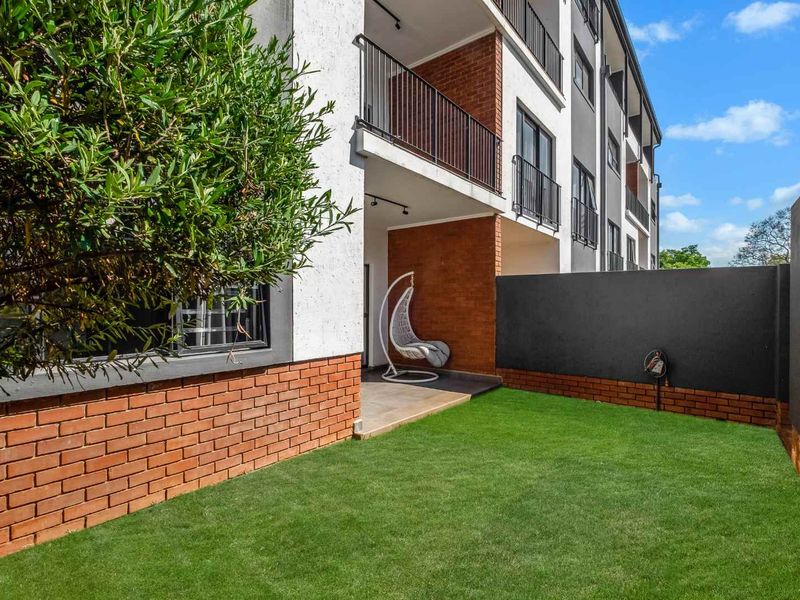 A private rear garden area featuring a covered patio, brick boundary walls, and well-maintained artificial lawn, attached to a modern apartment building.