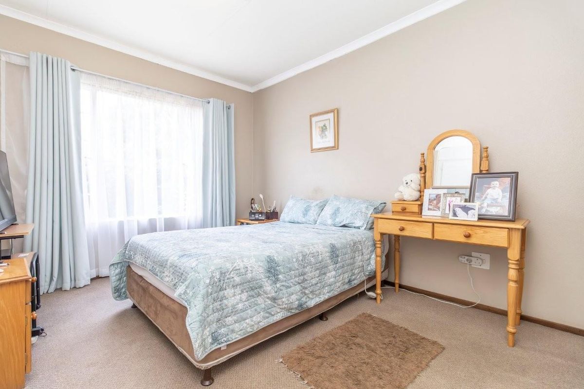 A well-lit bedroom featuring neutral wall tones, carpeted flooring, and large windows with fitted curtain tracks.