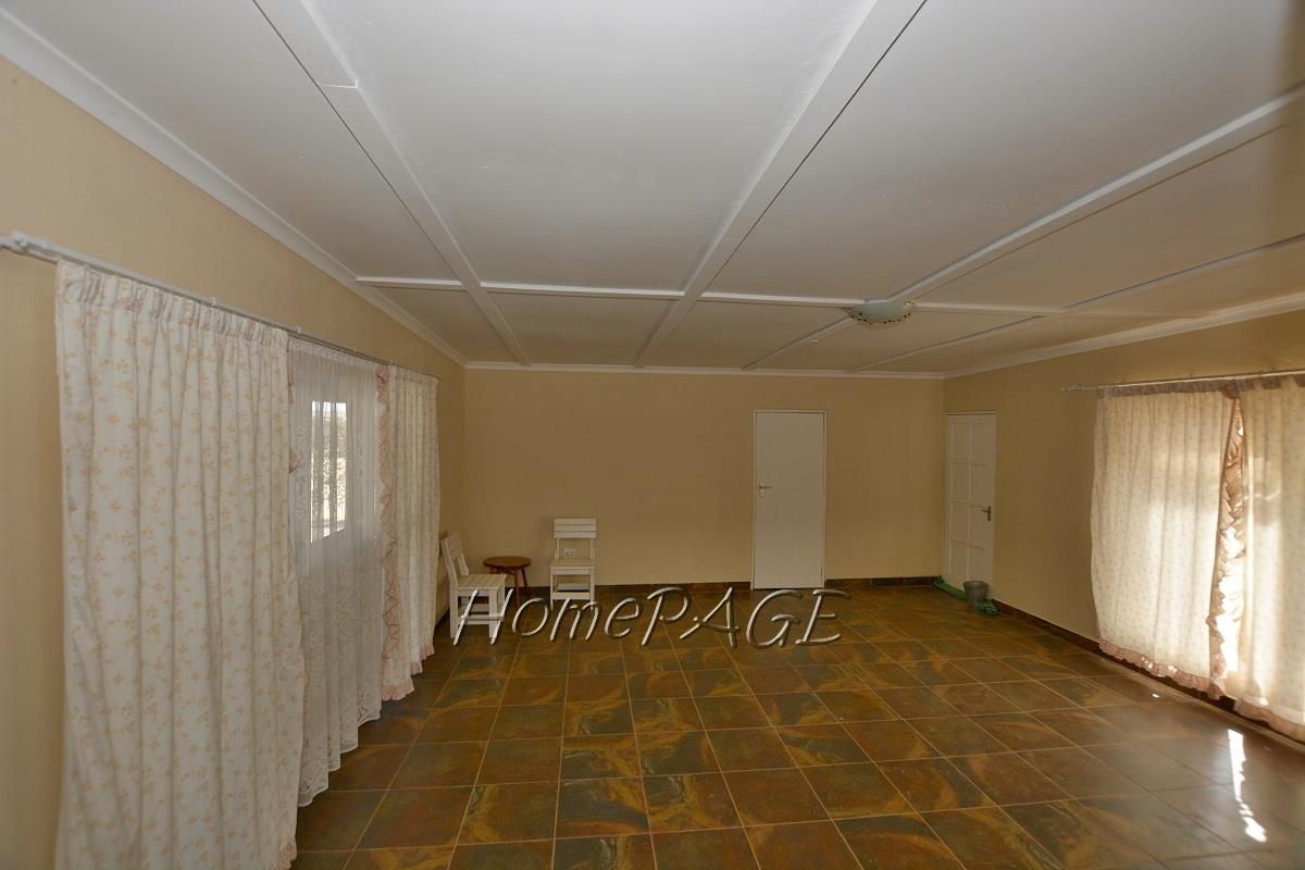 A spacious, empty living room featuring tiled flooring, a panelled ceiling, and multiple windows with curtain fixtures.