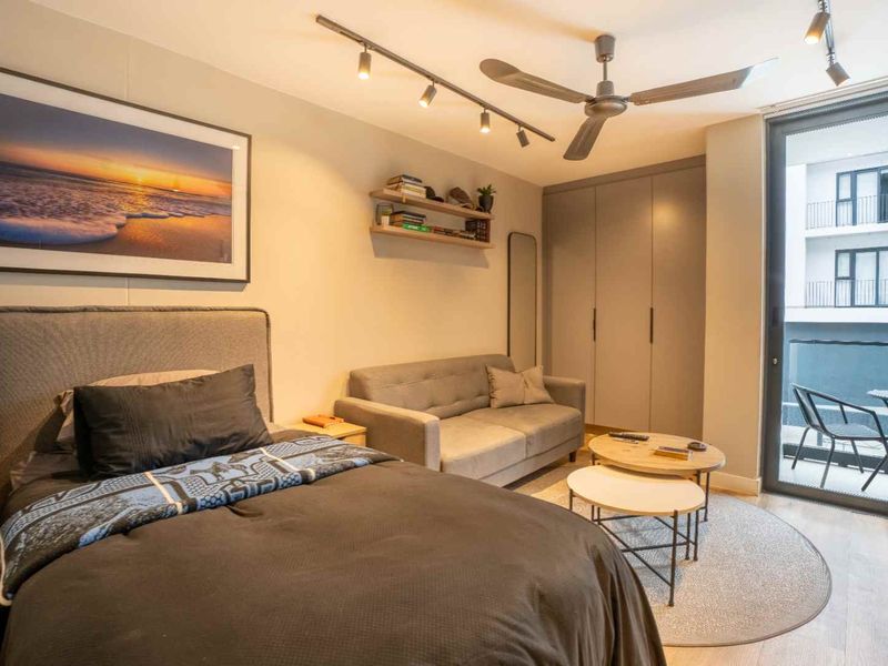 A well-maintained bedroom featuring built-in wardrobes, modern track lighting, a ceiling fan, and direct access to a private balcony through sliding glass doors.