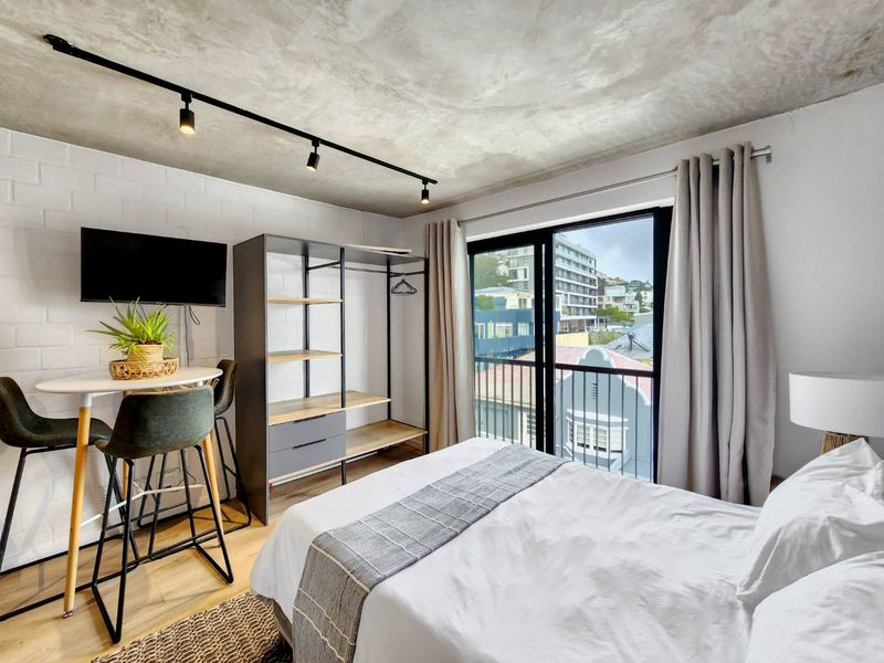 Modern bedroom featuring an exposed concrete ceiling, painted brick wall, and sliding glass doors leading to a balcony with urban views.