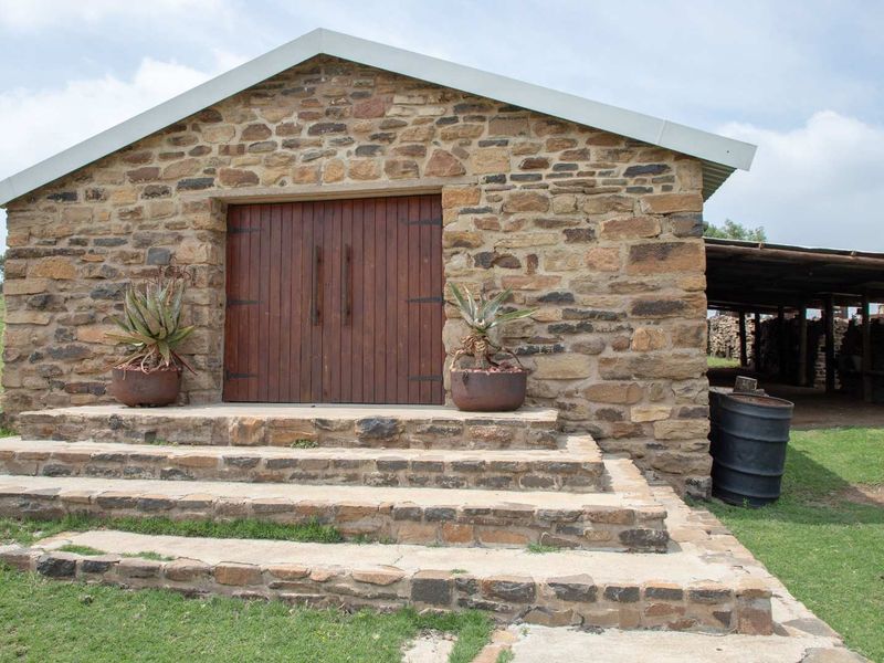 A rustic stone-built farm building featuring double timber doors, stone steps, and an adjacent covered storage area, set within a rural landscape.