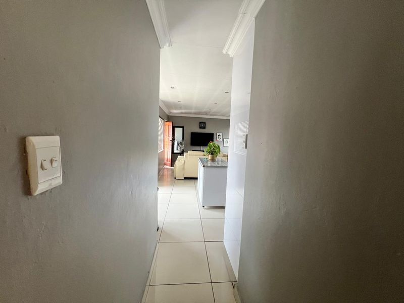 A view through a neutral-toned hallway leading into an open-plan living area with tiled flooring and recessed ceiling lighting.