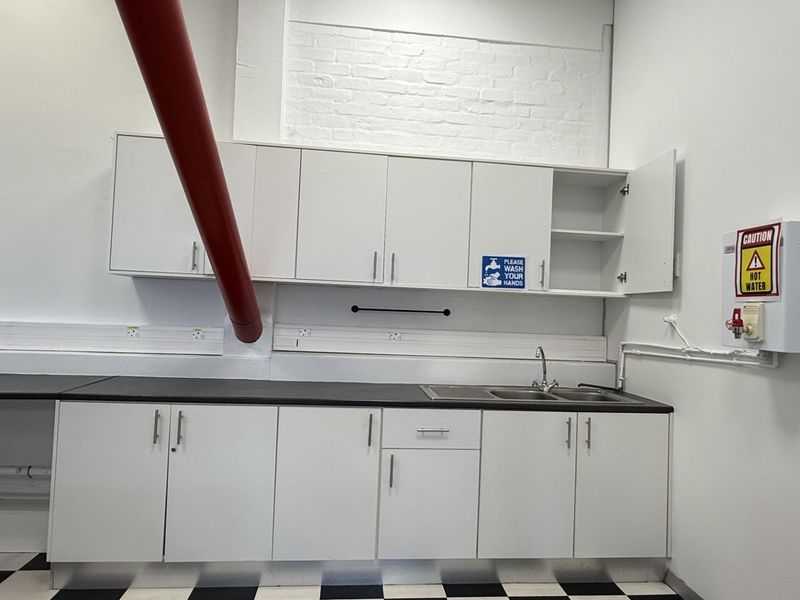 A functional office kitchenette featuring white cabinetry, a stainless steel sink, and a hot water dispenser, set against a clean, industrial-style backdrop.