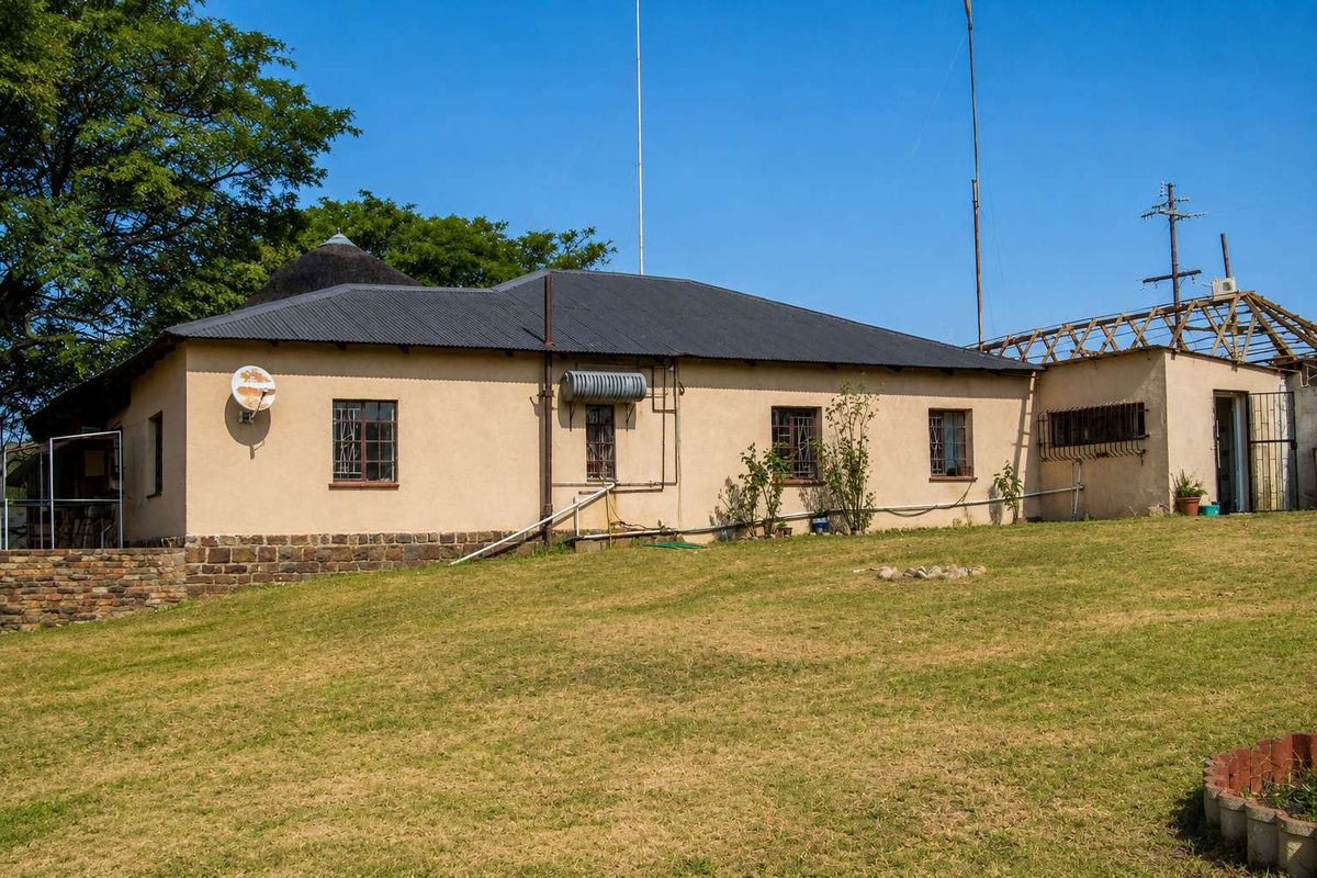Rear view of a farm-style dwelling featuring a corrugated metal roof, stone foundation, and security bars on windows, set against a large grassy yard.