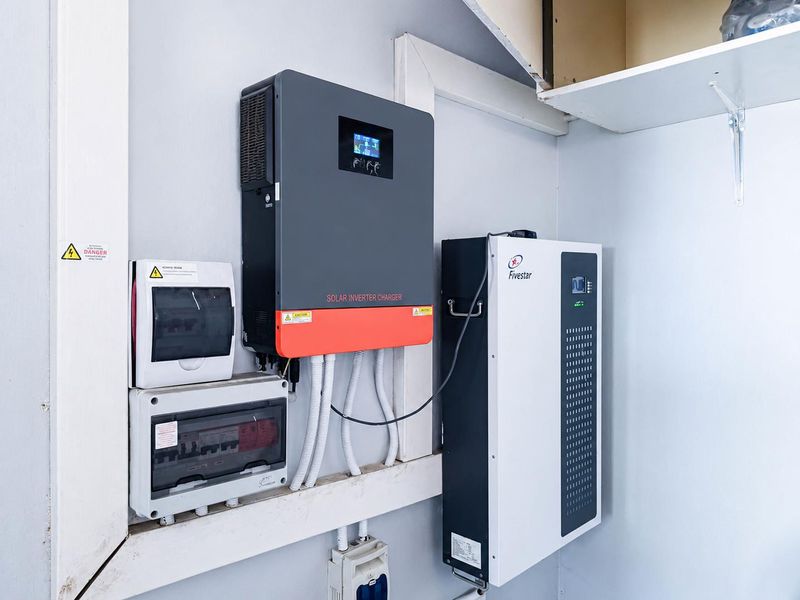 Utility area featuring a wall-mounted solar inverter charger, lithium battery storage unit, and electrical distribution board with circuit breakers.