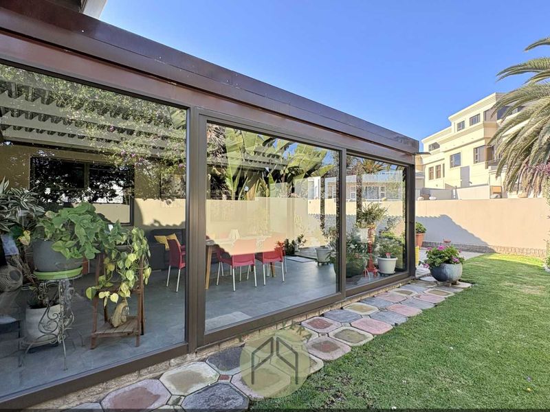 A well-maintained enclosed patio area featuring large glass panels, overlooking a landscaped garden with a paved stone walkway.