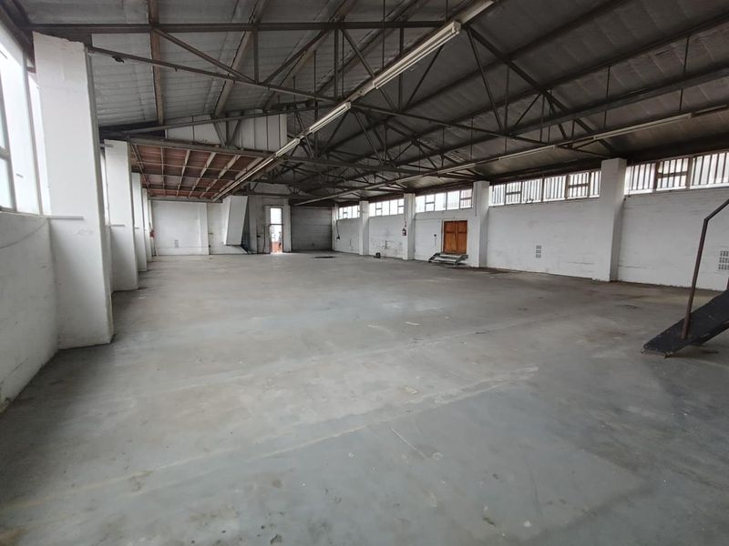 Spacious industrial warehouse area featuring high ceilings with exposed steel trusses, concrete flooring, and ample natural light through large windows.