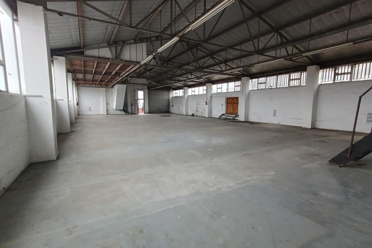 Spacious industrial warehouse area featuring high ceilings with exposed steel trusses, concrete flooring, and ample natural light through large windows.