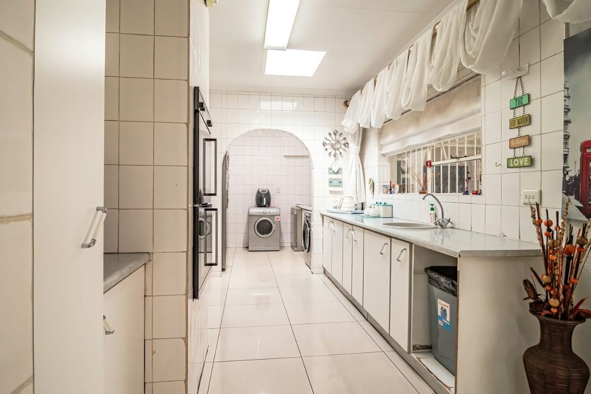 A dated kitchen featuring tiled walls and flooring, built-in cabinetry, and a stainless steel sink, with an adjacent laundry area visible through an arched doorway.