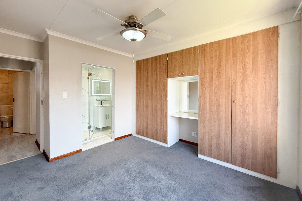 A bedroom featuring built-in wooden cupboards with a vanity area, a ceiling fan, and an en-suite bathroom with a glass-enclosed shower.