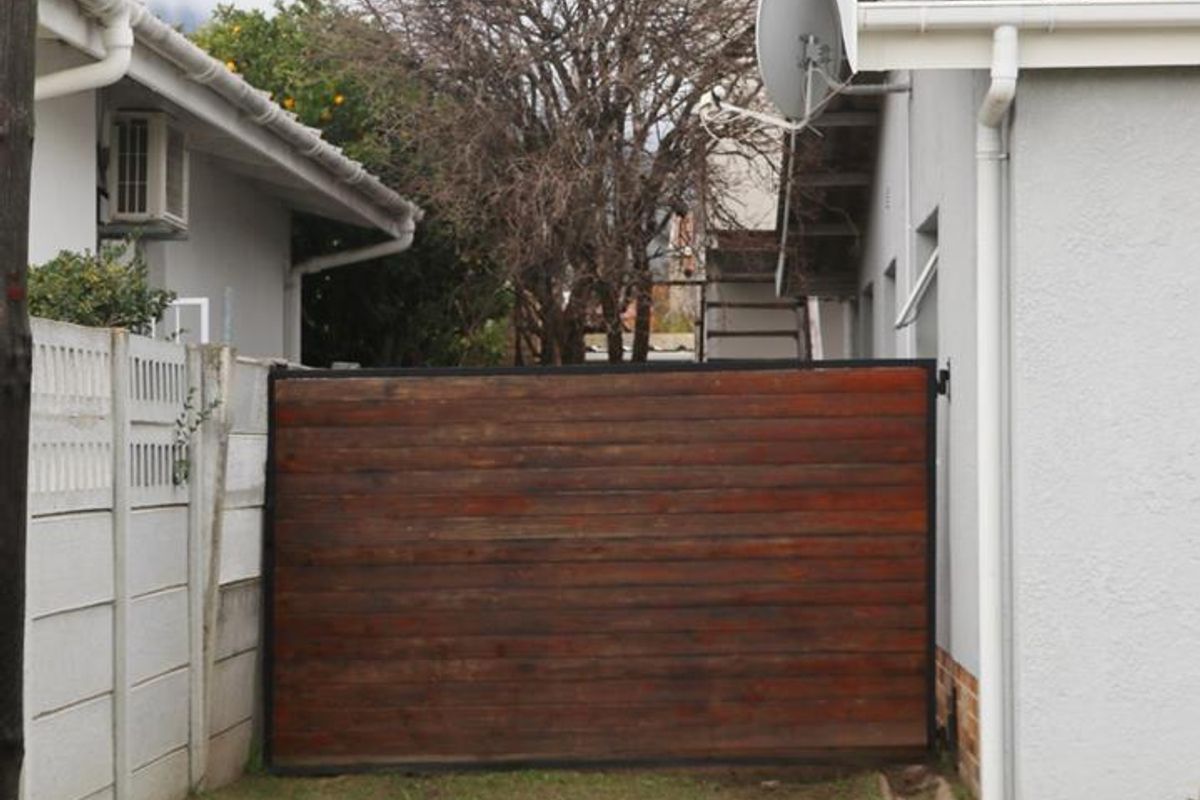 A rear exterior view showing a timber gate, precast concrete boundary wall, and drainage infrastructure between two residential structures.