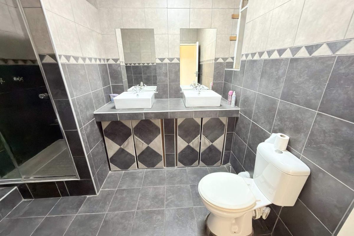 A functional bathroom featuring a double vanity with vessel sinks, a toilet, and a shower enclosure with grey and white tiled finishes.