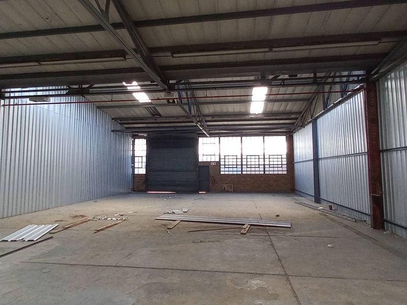 Spacious industrial warehouse unit featuring high ceilings with exposed steel trusses, concrete flooring, corrugated metal walls, and a large roller shutter door.