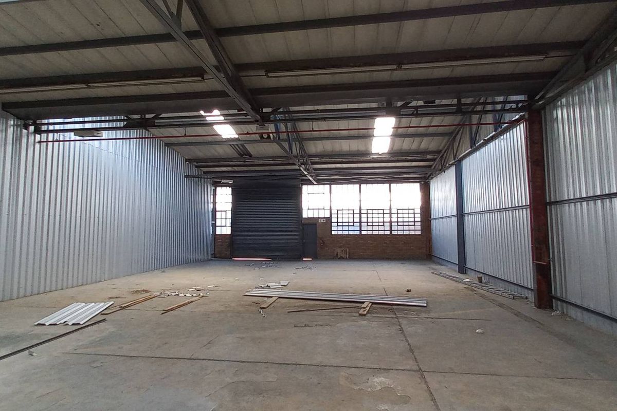 Spacious industrial warehouse unit featuring high ceilings with exposed steel trusses, concrete flooring, corrugated metal walls, and a large roller shutter door.