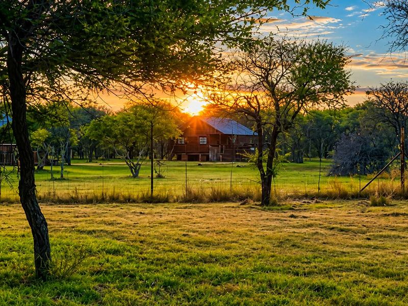 A scenic view of timber-clad farm buildings set within a lush, grassy landscape at sunset, featuring elevated decks and mature trees.