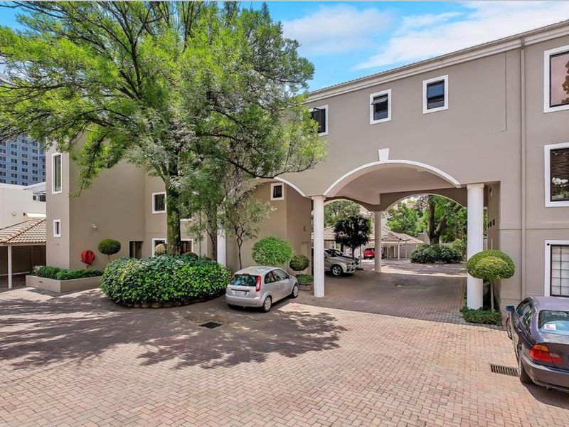 A well-maintained apartment complex entrance featuring a paved driveway, arched portico with columns, and mature landscaping.