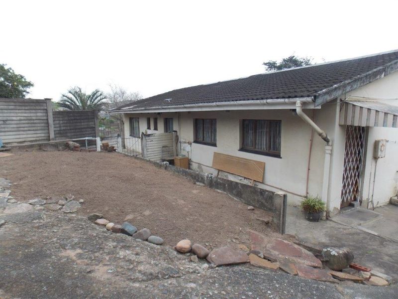 Rear exterior view of the house showing a paved area, concrete boundary wall, and basic building structure requiring maintenance and landscaping.