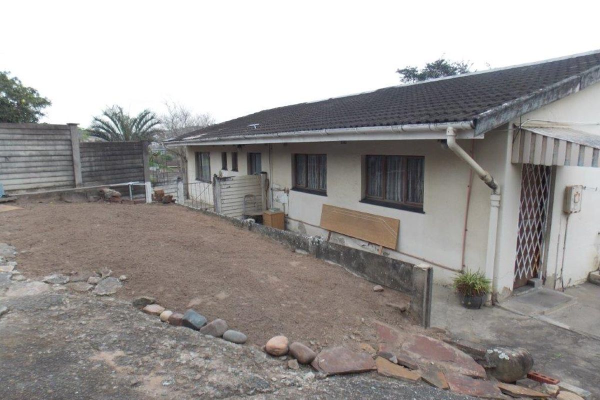 Rear exterior view of the house showing a paved area, concrete boundary wall, and basic building structure requiring maintenance and landscaping.