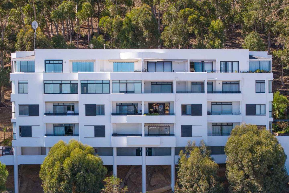 A multi-story apartment building facade featuring multiple balconies, large windows, and sliding glass doors, set against a wooded hillside.