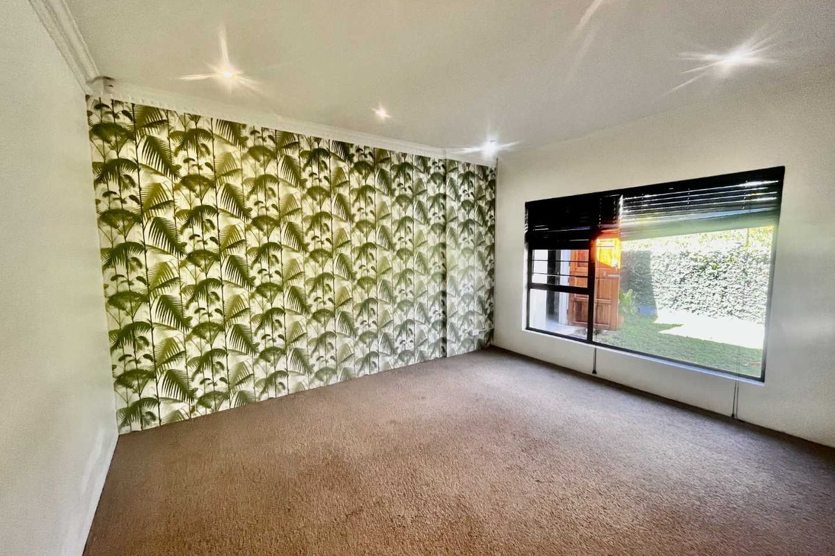 A bright, empty bedroom featuring a large window with blinds, recessed ceiling lighting, and a prominent palm-patterned feature wall.