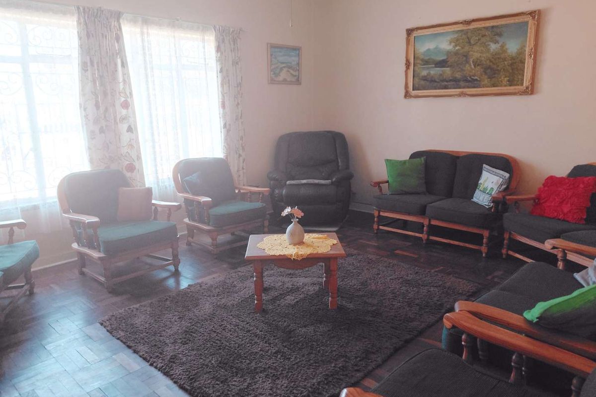 A traditional living room featuring classic parquet flooring and large windows fitted with security bars, offering a dated but well-maintained living space.
