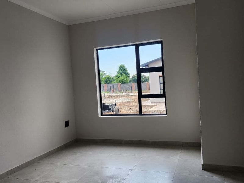 A bright, empty bedroom featuring neutral tiled flooring and a modern black aluminium window frame, offering a clean and versatile space.