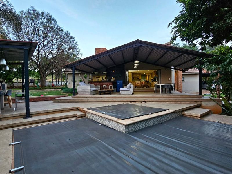 Spacious covered rear patio featuring a built-in braai area, overlooking a swimming pool with a safety cover and landscaped garden.