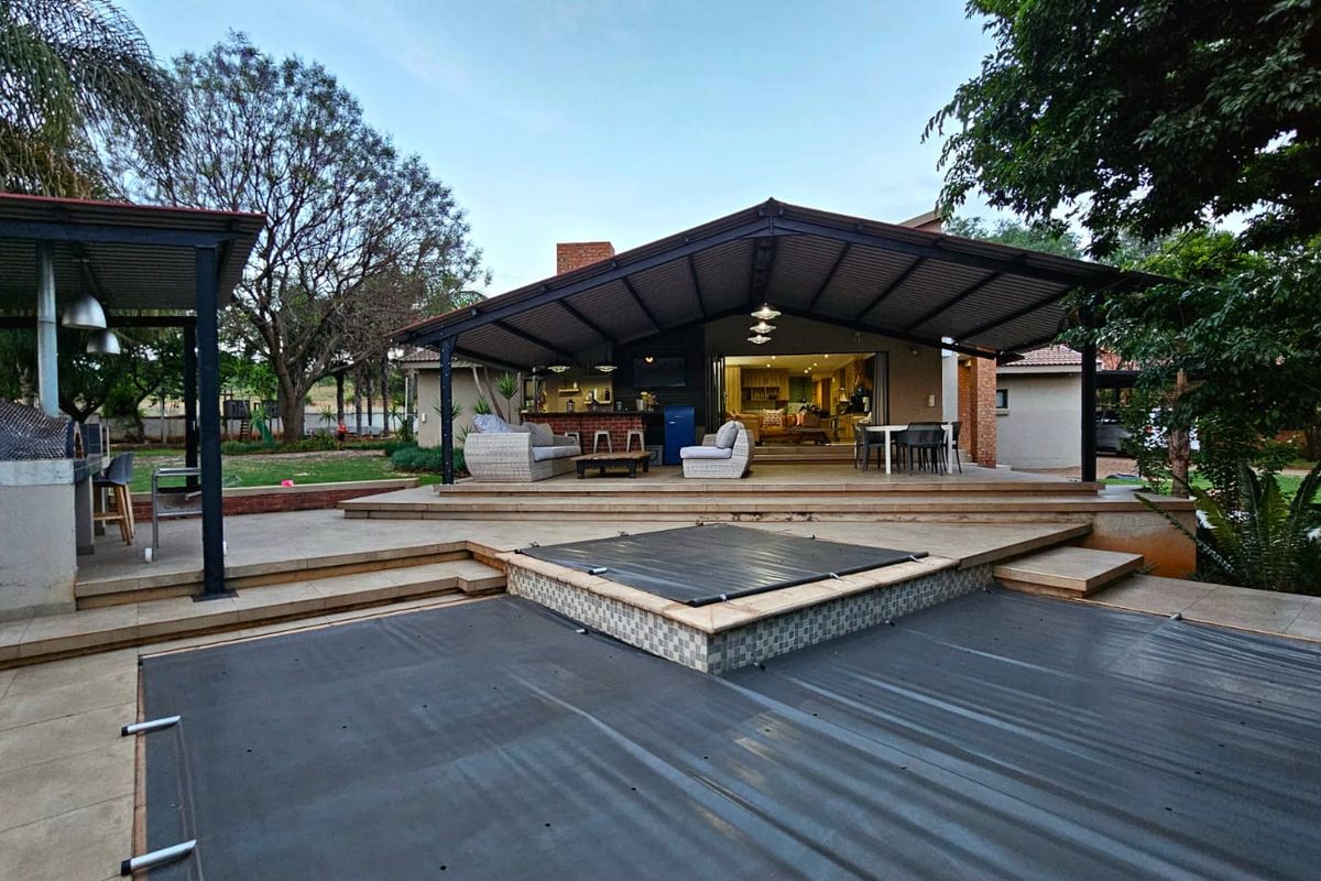 Spacious covered rear patio featuring a built-in braai area, overlooking a swimming pool with a safety cover and landscaped garden.
