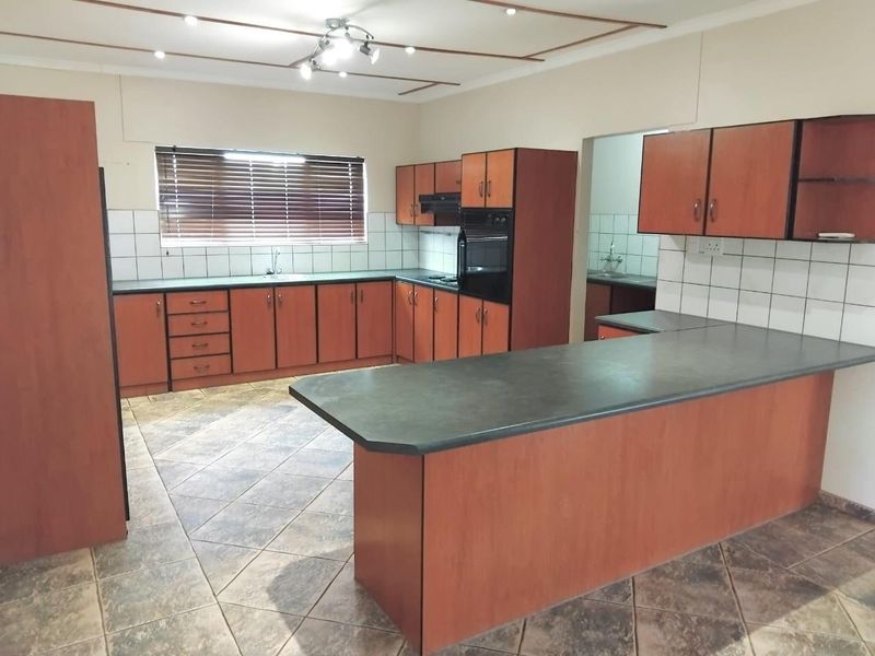 A spacious kitchen featuring wood-finish cabinetry, dark laminate countertops, a breakfast bar, and tiled flooring, with an adjacent scullery area visible.