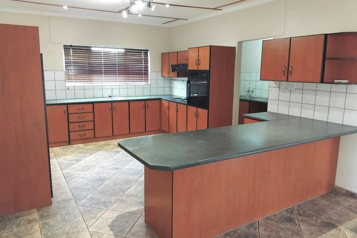 A spacious kitchen featuring wood-finish cabinetry, dark laminate countertops, a breakfast bar, and tiled flooring, with an adjacent scullery area visible.